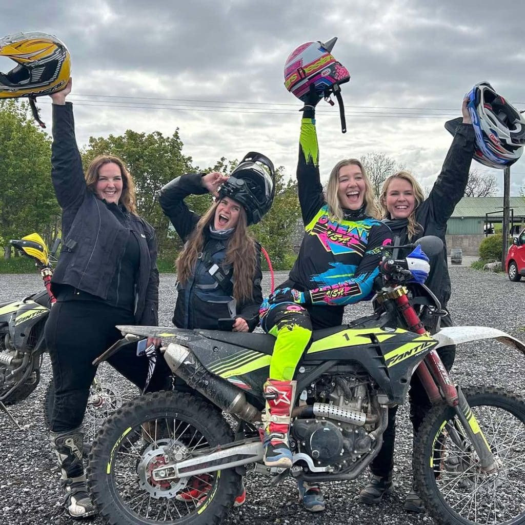 A small group of 4 women grinning and holding their dirt bike helmets in the air behind a trail riding motorcycle at the beginning of their dirt bike training day.